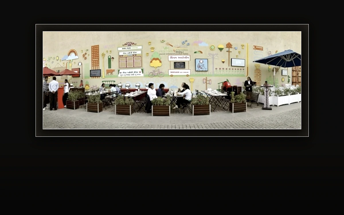 Open-air festive dining in front of a beige mural wall of illustrated shopfronts and Gujarati signage for Bhel Puri Centre and Shivam Ice Cream, with red and navy umbrellas, planters and diners
