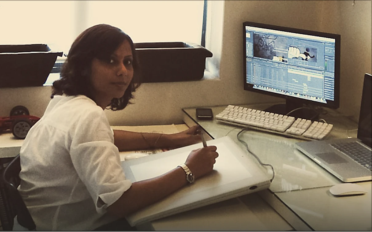 A designer at her studio workstation — stylus in hand over an angled drawing tablet, with a monitor running animation software behind